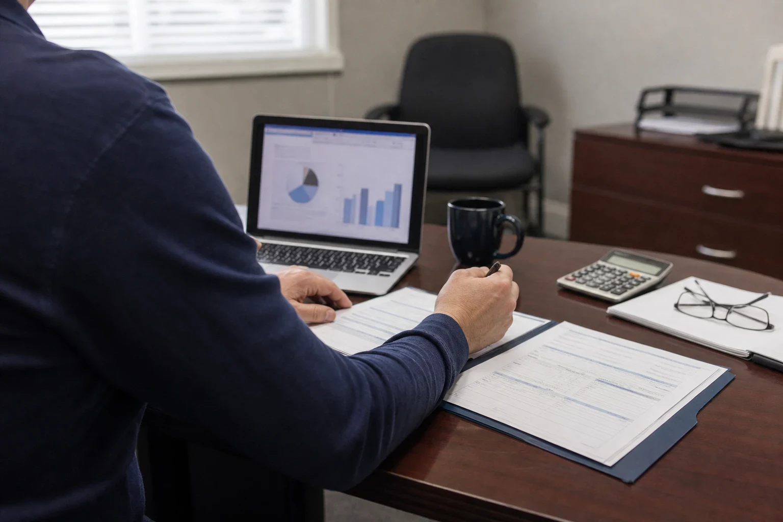 Over-the-shoulder view of a person in a navy sweater reviewing forms at a wooden office desk beside a laptop showing charts, a calculator, mug and reading glasses