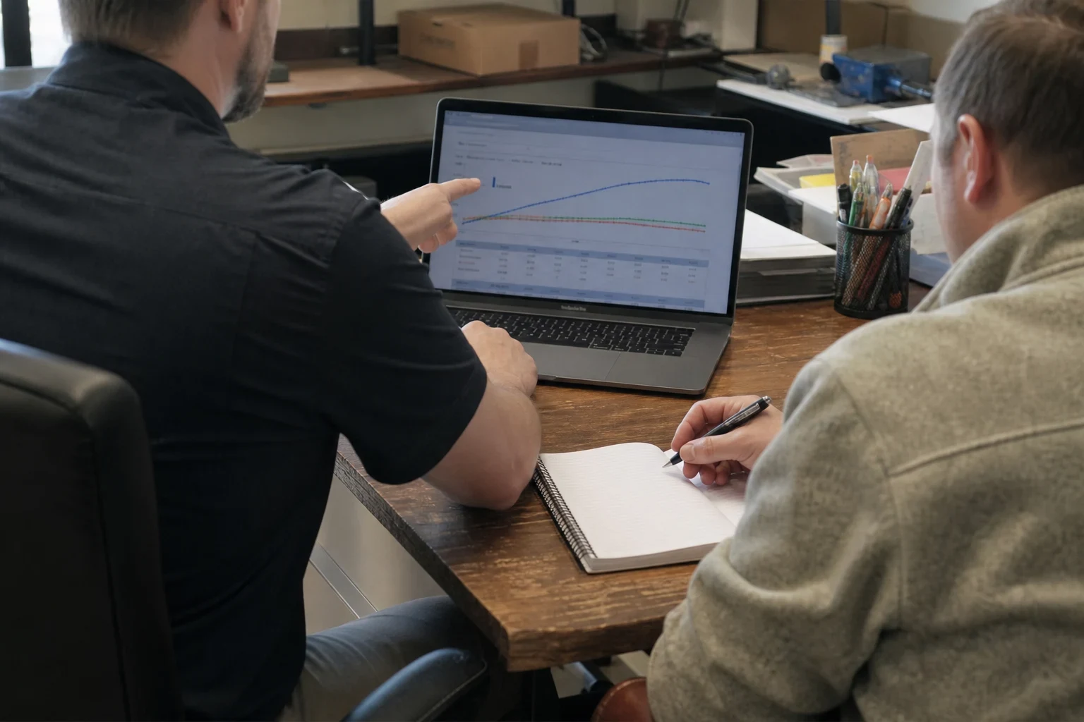Two men at a wooden office desk; one points to a line chart on a laptop screen while the other takes notes in a spiral notebook beside a cup of pens.